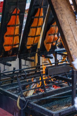 Kalın somon filetoları, Macaristan 'da Aralık 2025' te Budapeşte Noel Pazarı 'nda, ön planda beyaz çelenk bulunan bir gıda tezgahındaki açık odun ateşinde dikey olarak pişirilir.