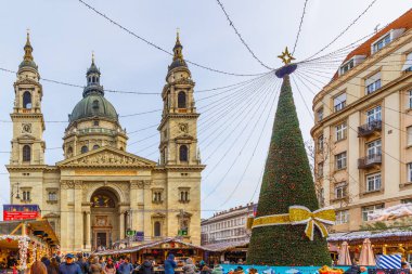 Turistler ve yerliler, Macaristan 'ın başkenti Budapeşte' de Aralık 2025 'te düzenlenen Advent Bazilika Noel Pazarı' nı demirleyen St. Stephen Bazilikası 'na doğru mevsimlik ışıklarla süslenmiş festival caddesi boyunca yürüyorlar.