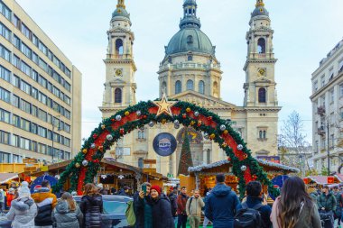 Turistler ve yerliler, Macaristan 'ın başkenti Budapeşte' de Aralık 2025 'te düzenlenen Advent Bazilika Noel Pazarı' nı demirleyen St. Stephen Bazilikası 'na doğru mevsimlik ışıklarla süslenmiş festival caddesi boyunca yürüyorlar.