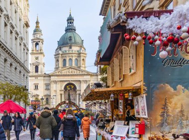 Turistler ve yerliler, Macaristan 'ın başkenti Budapeşte' de Aralık 2025 'te düzenlenen Advent Bazilika Noel Pazarı' nı demirleyen St. Stephen Bazilikası 'na doğru mevsimlik ışıklarla süslenmiş festival caddesi boyunca yürüyorlar.