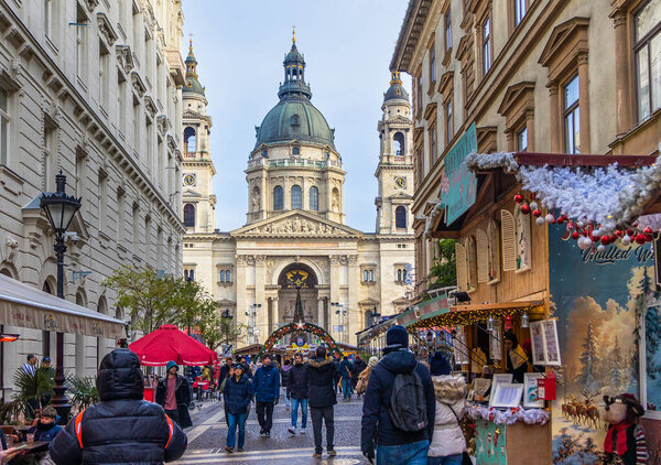 Туристы и местные жители идут по праздничной улице, украшенной сезонными огнями в сторону базилики Святого Стефана, которая якорь Рождественский рынок Адвент Базилика в Будапеште, Венгрия, декабрь 2025 года