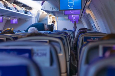 Koltukların arkasından çekilen yatay geniş açı açısı, ticari uçuş sırasında mavi multimedya ekranlı bir uçak kabinindeki insanları gösteriyor.