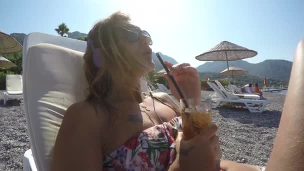 魅力的な女性がビーチで音楽とドリンク冷たいお飲み物をリッスンします。 — ストック動画