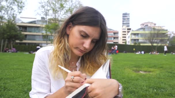 Ung kvinna skriver något på anteckningsboken i park — Stockvideo