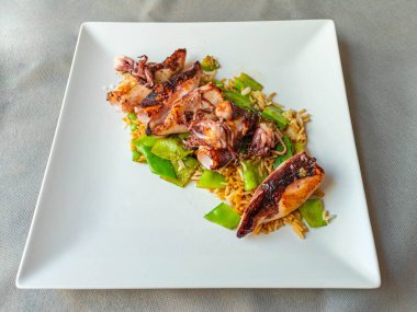 Kare beyaz bir tabakta servis edilen, ızgara kalamar ve taze bezelyeli pilav gurme tabağı. Deniz ürünleri, Asya mutfağı ve sağlıklı tarif konsepti..