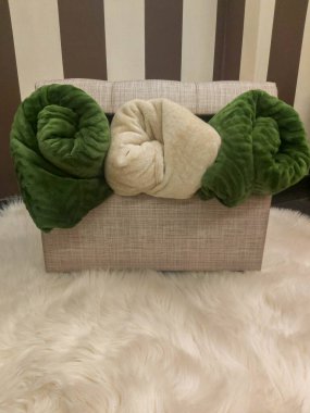 Beyaz bir kürk halısının üzerinde örülmüş sepetin içinde üç adet yeşil ve bej tüylü battaniyenin dikey çekimi. Ev konforu, rahat kış ve iç tasarım kavramı.