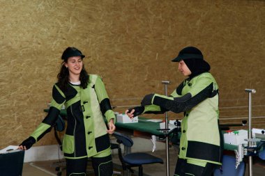 Atış teçhizatlı iki genç kadın atış talimine hazırlanıyor. Konferansta hassasiyet ve takım çalışması, rekabetçi spor ve disiplinli eğitim konuları ele alınıyor.