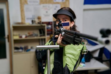 Bir kadın atış poligonuna tüfeği doğrultuyor, hassasiyet ve odaklanma gösteriyor. Bu resim rekabetçi spor, hedef çalışması veya doğruluk ve beceri arayışı için uygundur..