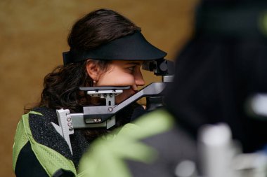 Genç bir kadın, bir atış yarışması sırasında tüfeği nişan alırken dikkatle odaklanır. Bu görüntü, hedef atışı için gereken hassasiyeti, yeteneği ve konsantrasyonu yakalar.