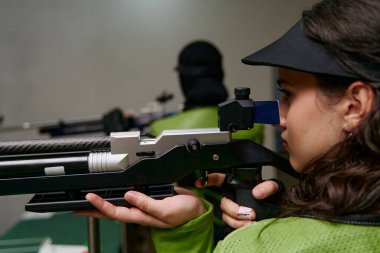 Genç bir kadın havalı tüfekle kapalı bir atış poligonuna nişan alıyor. Hassasiyet ve odaklanma gösteriyor. Bu resim rekabetçi atış, hedef çalışması ve profesyonel atış için uygundur.