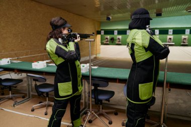 Atış poligonunda iki bayan sporcu havalı tüfekle pratik yapıyor. Hedefe nişan alıyorlar. Odaklanma ve hassasiyet gösteriyorlar. Bu görüntü, içerdiği yoğunluğu ve beceriyi yakalar.