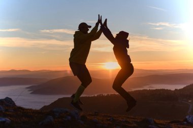 Çift gün batımında bir dağın zirvesinde havaya sıçrayıp renkli bir gökyüzüne karşı siluetler oluşturuyor. Sahne, doğada neşe ve dostluğu yakalar. Güzellik..