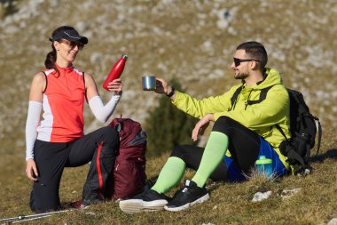 İki arkadaş içmek ve rahatlamak için dağ yolunda mola verir. Gülüyorlar ve güneşli bir günde doğa tarafından çevrelenmiş açık hava kıyafetleri giyiyorlar..