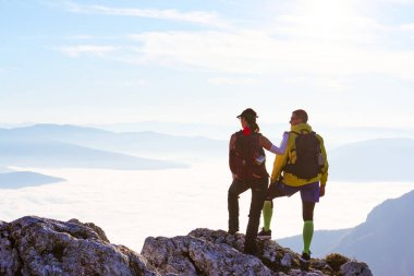 İki yürüyüşçü kayalık bir zirvede durup aşağıdaki büyüleyici manzaraya bakıyorlar. Güneş uzaklardan batıyor, vadiyi kaplayan bulutların üzerine sıcak bir parıltı saçıyor. Çift nefes kesici manzaranın keyfini çıkarıyor.