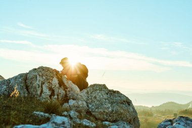Bir insan gün batımında dağlık bir alanda engebeli kayaların üzerinden tırmanıyor. Güneş gökyüzüne karşı parıldayan bir siluet yaratıyor. Yürüyüşün doğal güzelliğini ve macerasını vurguluyor..