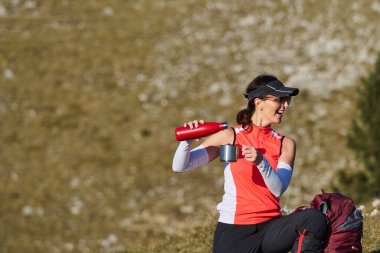 Parlak kıyafetli bir kadın dağlarda yürüyüş yapmaya ara verir ve gülümserken bardağa sıcak bir içecek döker. Güneşli bir günde yemyeşil tepeler etrafını sarar.