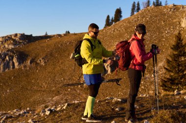 Yürüyüşçüler dağ maceralarına hazırlanıyorlar. Güneş batarken teçhizat ve sırt çantalarını ayarlıyorlar. Engebeli manzara, canlı giysilerini ve yürüyüş ekipmanlarını vurguluyor..