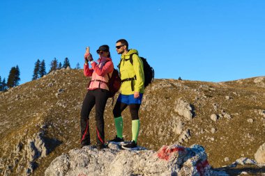 İki yürüyüşçü dağdaki kayalık bir çıkıntıda fotoğraf çekmek için durdular. Parlak mavi gökyüzü ve altın manzara maceraları için manzaralı bir zemin oluşturuyor..