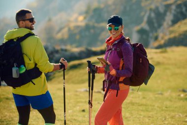 İki yürüyüşçü dağlardaki açık hava maceraları sırasında gülümseyip etkileşime geçiyorlar. Parlak güneş ışığı ve yeşil manzara birlikte doğayı keşfederken canlı bir atmosfer yaratır..