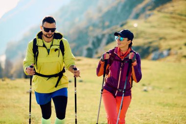 İki yürüyüşçü güneşli bir günde canlı yeşil bir çayırda yürürler, doğa ve temiz havanın tadını çıkarırlar, açık hava aktiviteleri için uygun renkli spor kıyafetleri giyerler..