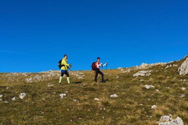 İki arkadaş çimenli bir arazide kaya çıkıntılarıyla yürüyüş yapıyor. Yürüyen sırıklar taşırlar ve güzel bir dağ manzarasında güneşli havanın tadını çıkarırlar..