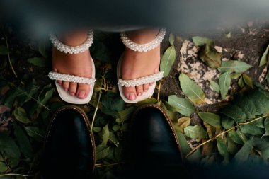 Sonbahar yapraklarıyla kaplı bir zeminde birbirine bakan bir erkekle bir kadının ayaklarını gösteren bir görüntü. Kadın incilerle süslenmiş zarif beyaz sandaletler giyer, erkek ise...
