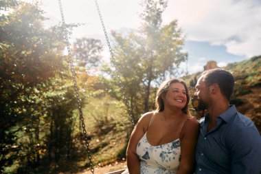 Mutlu çift doğada salıncakta romantik bir anın tadını çıkarıyor. Sahne, doğal güneş ışığıyla aydınlanıyor. Yumuşak bir mercek parıldıyor. Duygusal bağlantılarını ve samimiyetlerini vurguluyor..