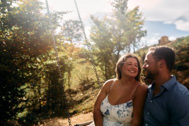 Mutlu çift doğada salıncakta romantik bir anın tadını çıkarıyor. Sahne, doğal güneş ışığıyla aydınlanıyor. Yumuşak bir mercek parıldıyor. Duygusal bağlantılarını ve samimiyetlerini vurguluyor..