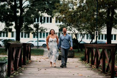 Neşeli bir genç adam ve kadın, yeşil bir şehir parkında kırsal ahşap bir yaya köprüsünde yürürken el ele tutuşarak romantik bir gezinti yapıyorlar..