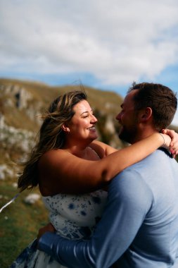 Bir erkeği kucaklarken sıcacık gülümseyen genç bir kadının yakın plan portresi. Çift, parlak bir gökyüzünün altında manzaralı, çimenli bir alanda birlikte bir anın tadını çıkarıyor..