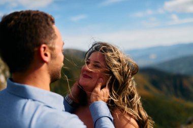 Dağlara yapılan bir gezi sırasında samimi bir anı paylaşan bir çiftin samimi bir görüntüsü. Kadın mutlu, otantik bir tebessümle ışıldarken, erkek sevgiyle sarılır kadının yüzüne.