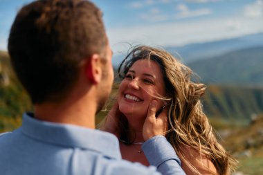 Dağlara yapılan bir gezi sırasında samimi bir anı paylaşan bir çiftin samimi bir görüntüsü. Kadın mutlu, otantik bir tebessümle ışıldarken, erkek sevgiyle sarılır kadının yüzüne.