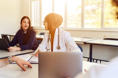 Başörtüsü takan bir tıp uzmanı, parlak bir sınıfta bayan bir öğrenciyle konuşuyor. Meslektaşları notlara, öğrenme, işbirliği ve hastalara işaret ederken dizüstü bilgisayar kullanıyorlar.