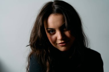 Uzun siyah saçlı, ince makyajlı ve karamsar ışıklandırmalı düşünceli bir kadının yakın plan portresi. Minimalist arka plan, yaşam tarzı, güzellik ve moda için mükemmel olan yüz ifadesini vurguluyor.