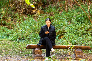 Gecelikli ve kot pantolonlu Asyalı bir kadın yeşil bahçede ya da parkta otururken ağzı kapalı telefona gülüyor. Dinleniyorum, dışarıda dinleniyorum. Yüksek kalite fotoğraf