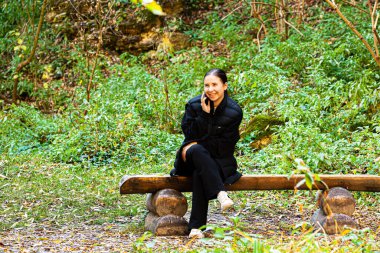 Gecelikli ve kot pantolonlu Asyalı bir kadın yeşil bir bahçede ya da parkta koltukta otururken telefonla konuşuyor. Dinleniyorum, dışarıda dinleniyorum. Yüksek kalite fotoğraf