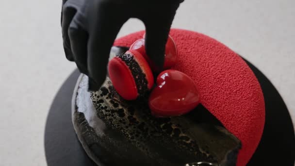 Confeiteiro decorando bolo de revestimento de veludo com elementos de sobremesa em forma de coração e macaron vermelho. Bolo para o dia dos namorados. Bolo de cor preta e vermelha — Vídeo de Stock