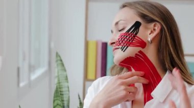 Boynunda ve yanağında kırışıklıklar olan Kinesio bantlı güzel bir kadın pencereye ve kameraya bakar. Yüz gerdirme güzellik prosedürü. Kineziyoloji tedavisi yakın çekim
