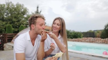 Young woman feeding her laughing friend a slice of cheesy pizza near poolside. Couple eating pizza together