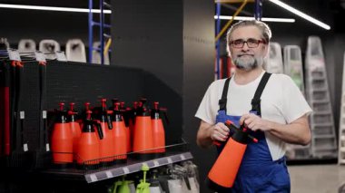 İş üniformalı bir adam mağazada elinde sprey şişeyle duruyor. Etrafı çeşitli alet edevat ve ekipmanlarla dolu raflarla çevrili.