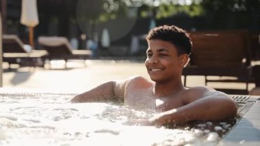 Genç siyah adam jakuzide yüzüyor, kabarcıklarla sıcak suda dinleniyor, hidroterapinin ve tatilin tadını çıkarıyor.