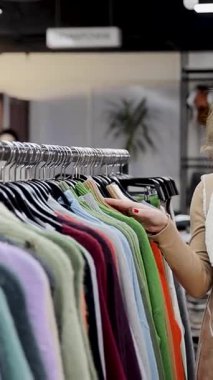 Elbise mağazasındaki askılarda asılı kalın süveteri seçen bir kadın. Kış kıyafetleri koleksiyonu. Sıcak giysi konsepti alıyorum. Dikey video