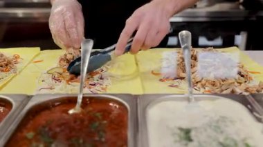 Restoran çalışanının elleri eldivenle çoklu shawarma paketleri hazırlıyor, tavuk, lahana ve turşu gibi malzemeler ekliyor.