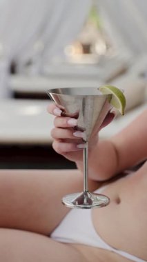 Beyaz bikinili bir kadın limon garnitürlü ferahlatıcı bir kokteyl tutuyor yaz otelinde lüks bir gündüz yatağında dinleniyor. Dikey video