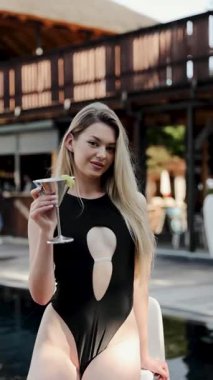 Siyah mayo giymiş genç bir adam havuz kenarında rahatlarken içki sergiliyor, kaygısız bir yaz havasının örneğini veriyor. Dikey video