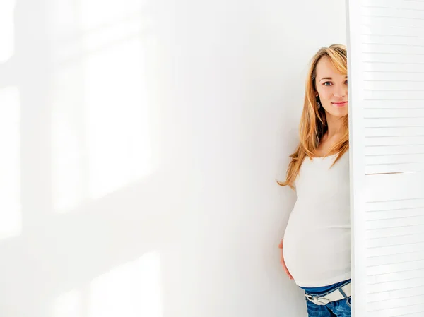 Fiatal és szép terhes nő úgy néz ki, egy fehér képernyő mögött a háttér fehér fal — Stock Fotó