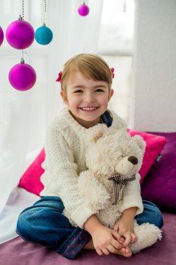 Güzel küçük bir kız bir pencere üzerinde oturan ve bir oyuncak ayı kucaklıyor. Pencere yeni yıl topları ile dekore edilmiştir.