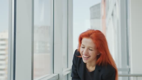 Женщина смотрит в камеру и смеется. Рыжеволосая девушка стоит у окна в офисе и тяжело смеется. Портрет счастливой женщины — стоковое видео