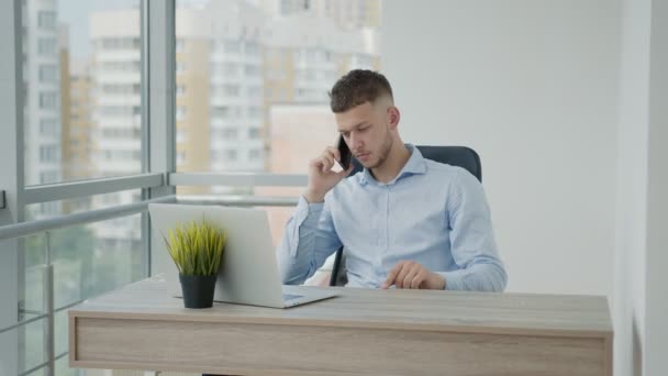 Mladý muž kavkazského vzezření emocionálně mluví do telefonu v kanceláři sedí u stolu. Vyřešit případy s mobilním telefonem a moderní gadgets. — Stock video