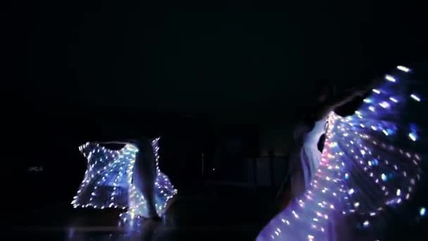 As meninas realizam uma dança com asas LED. Luzes LED brilham no escuro. Raparigas giram em torno de si mesmas e dançam no escuro. — Vídeo de Stock
